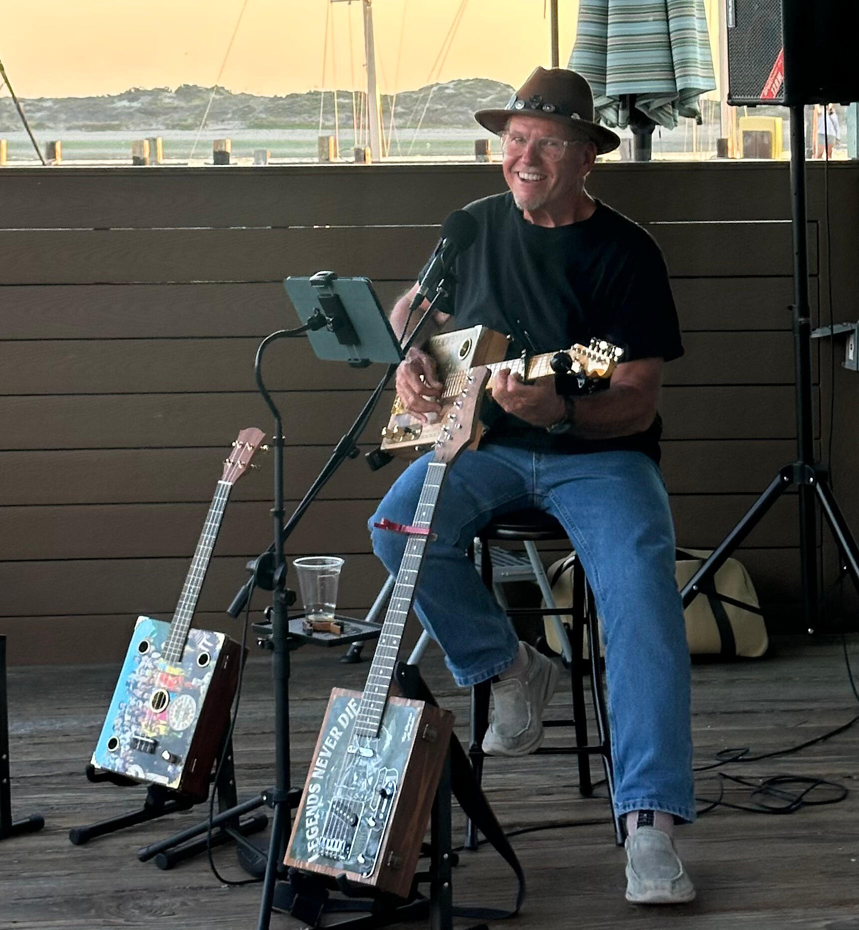Photograph of Ray Riordan playing one of his cigar box guitars in front of a brown fence with a market umbrella, sailboat masts, bay, and a sandbar behind the fence. At Ray's feet are two additional cigar box guitars on stands and a speaker on a stand to h